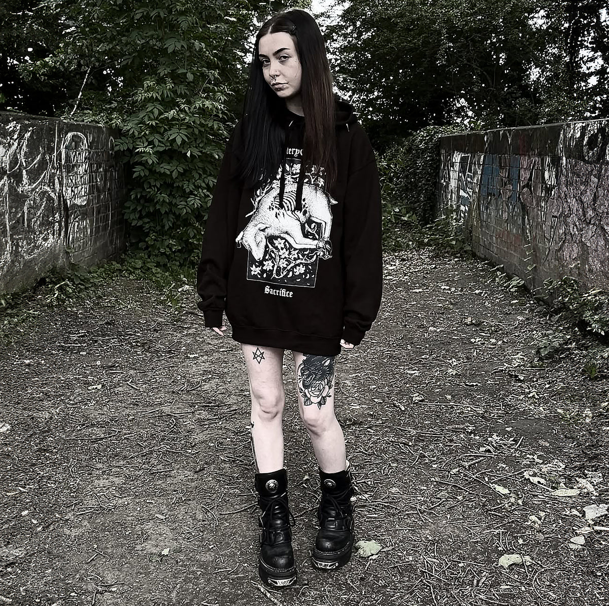 Model wearing the black Sacrificial Lamb Hoodie on an outdoor path beside a graffitied wall